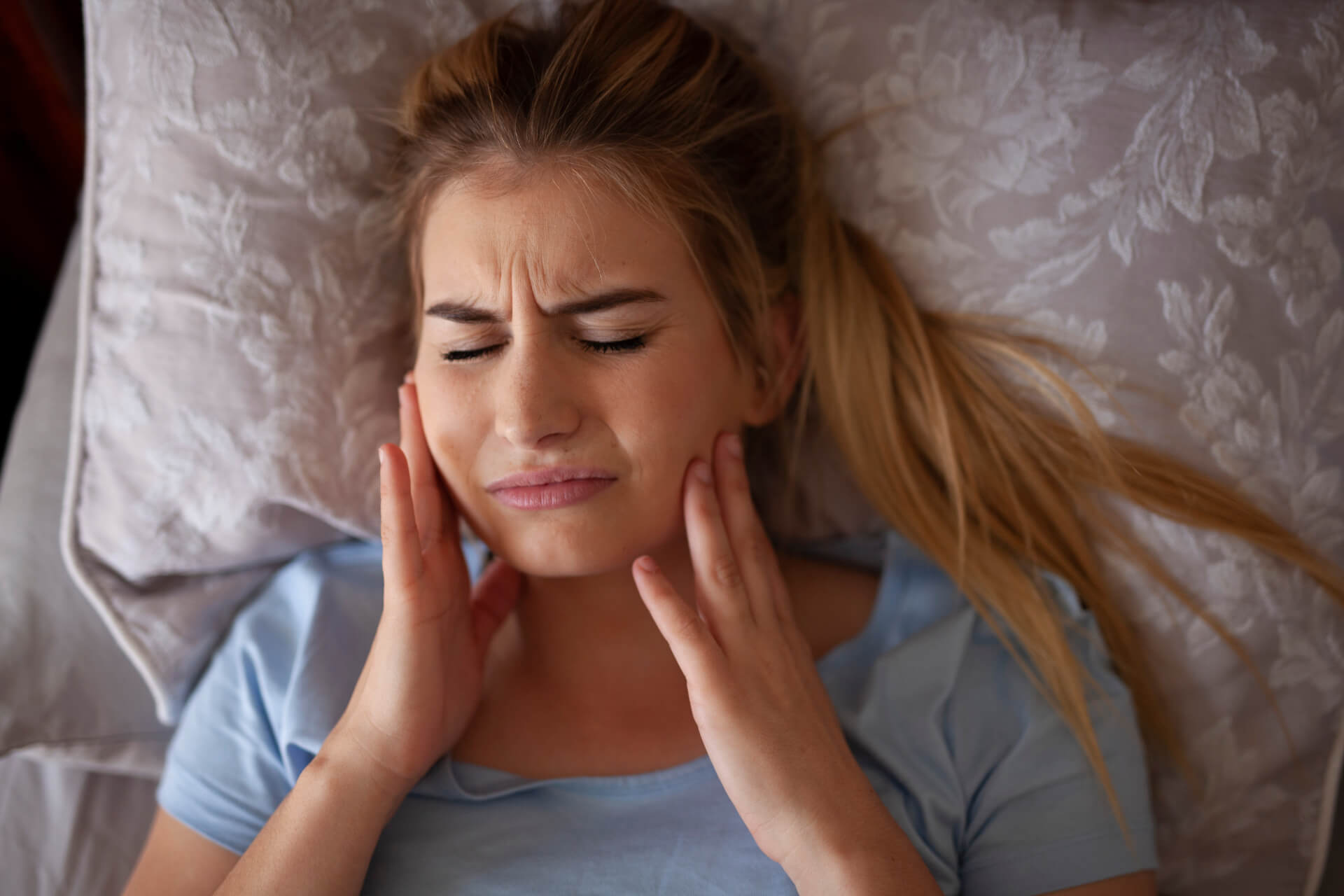 Woman laying in bed holding her jaw in pain.